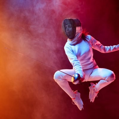 Blooming. Teen girl in fencing costume with sword in hand isolated on black background, neon lighted smoke. Practicing and training in motion, action. Copyspace. Sport, youth, healthy lifestyle.